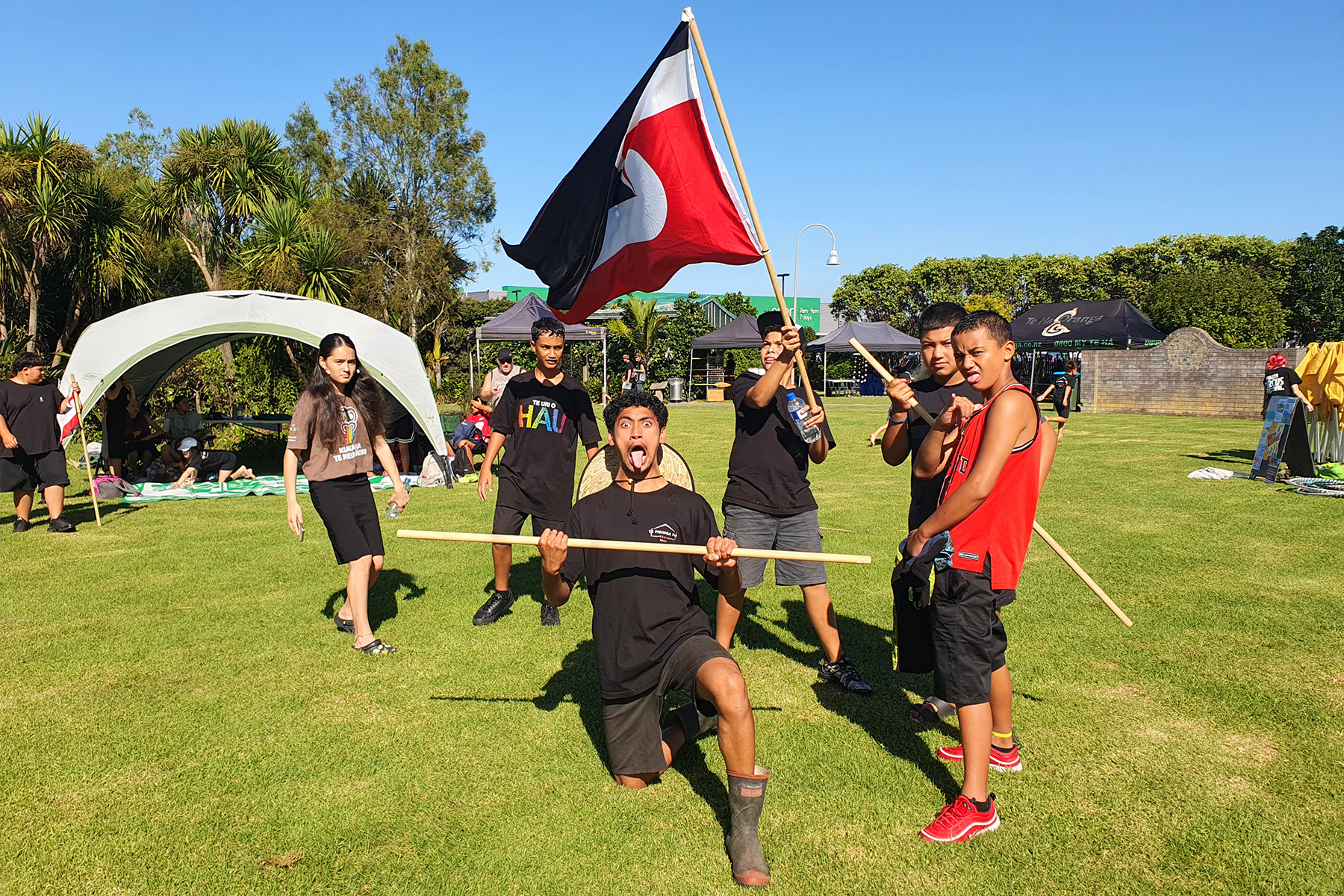 Six rangitahi pose in front of a Tino Rangitiratanga flag on grass. Tents, trees and a blue sky in the background.