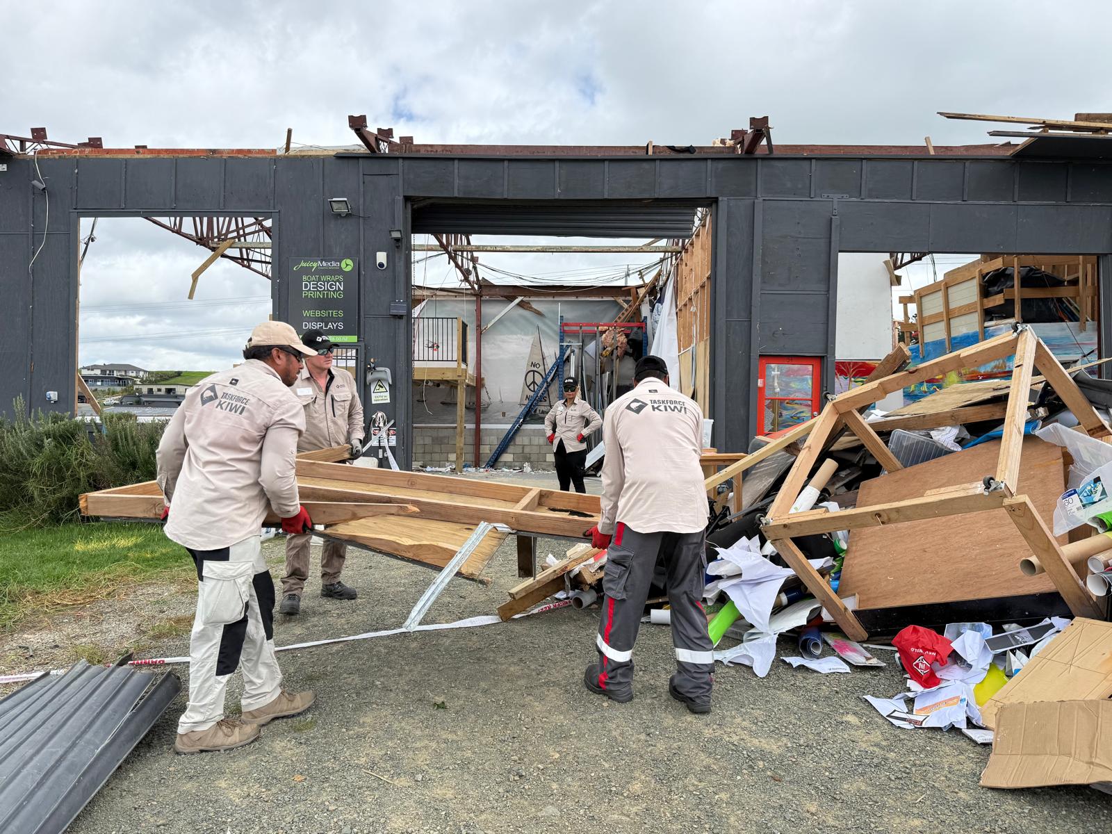 Workers in uniform remove building debris and rubble, moving it to a large pile. In the background, a partially demolished commercial shed.