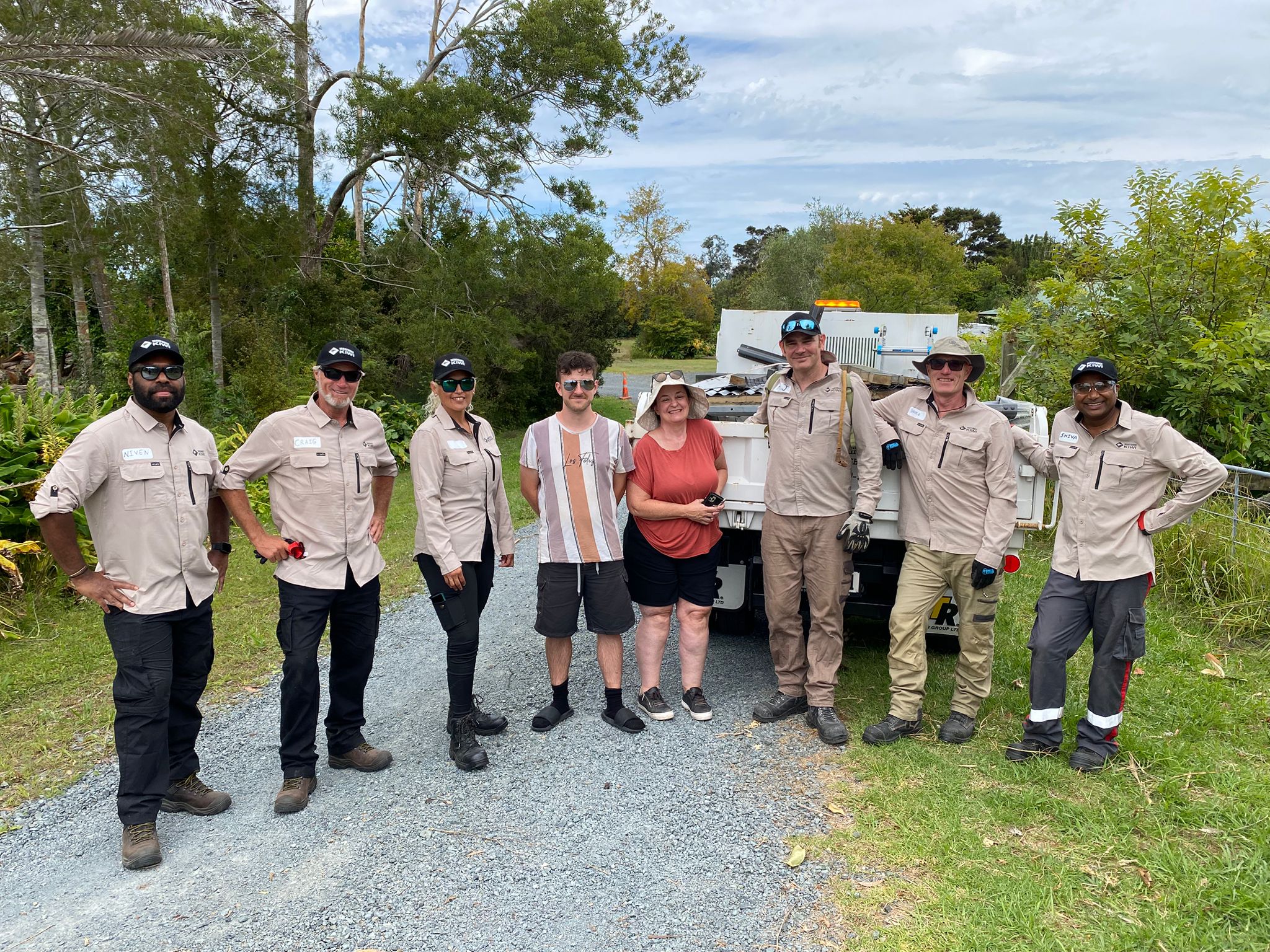 A group of people in uniform stand facing the camera and smiling, with a woman and a man. The people stand in front of a truck filled with building debris.